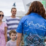A woman wearing a "Jellurgal Aboriginal Cultural Centre" shirt stands facing a family of three near a docked boat.