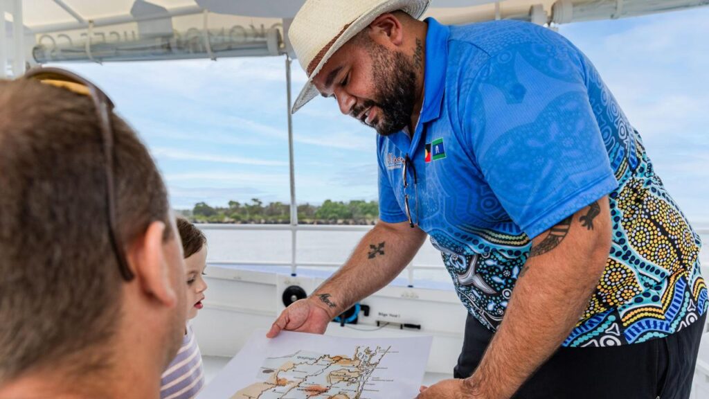 A man in a hat and patterned shirt shows a map to two people on a boat, with water and trees visible in the background.