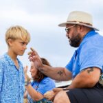 An adult in a patterned blue shirt applies ochre to a boy’s forehead near a beach, while another person sits in the background.
