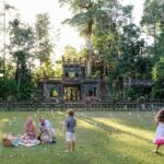 A family enjoys a picnic on grass as children play with bubbles in front of a historic stone structure surrounded by lush trees.