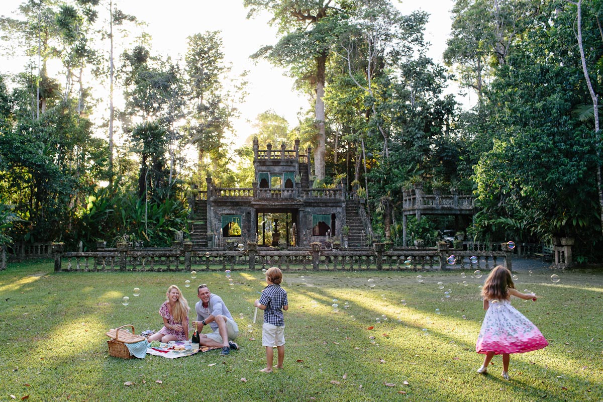 A family enjoys a picnic on grass as children play with bubbles in front of a historic stone structure surrounded by lush trees.