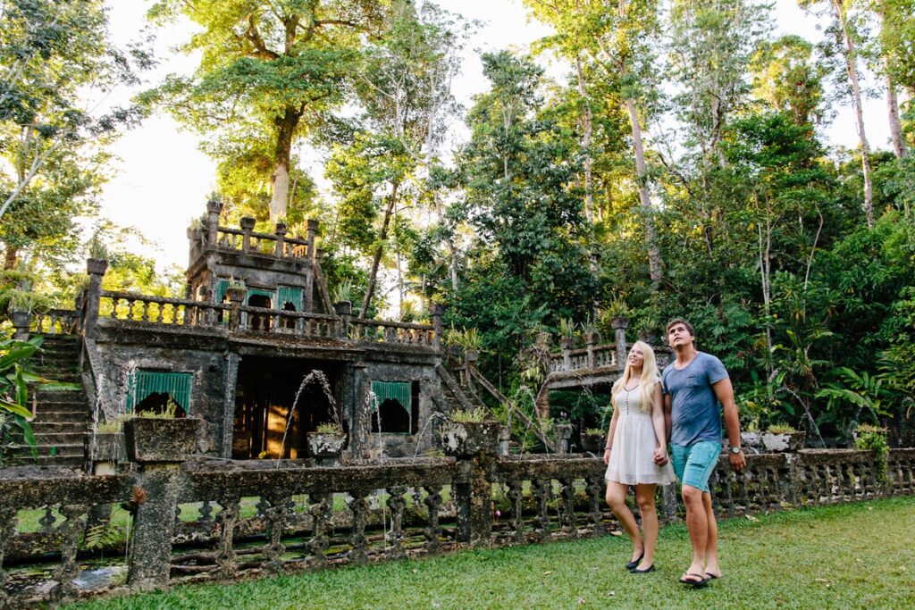 A couple walks hand in hand outside an old stone structure surrounded by lush greenery and trees.