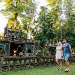 A couple walks hand in hand outside an old stone structure surrounded by lush greenery and trees.