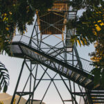 A tall metal observation tower with stairs, surrounded by trees, stands against a clear sky with mountains visible in the background.