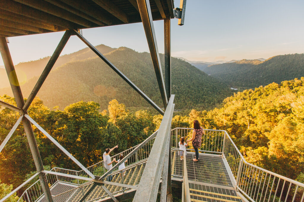 Three people stand on a metal observation platform overlooking a forested valley and distant mountains during sunset.
