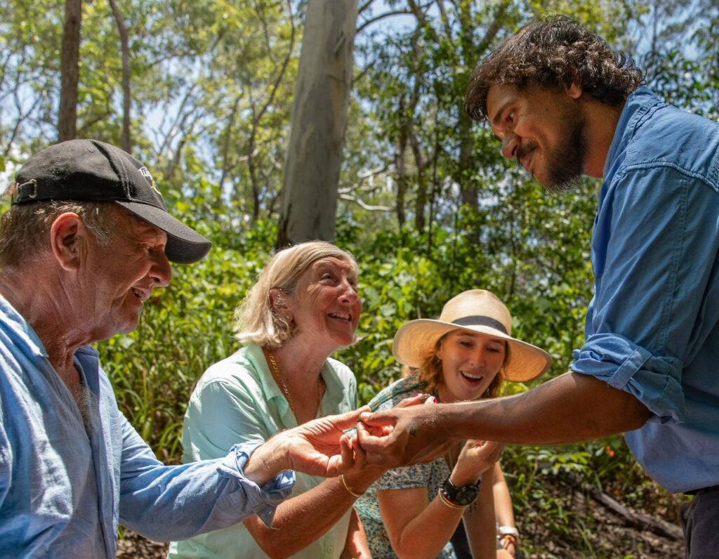Four adults outdoors in a forested area observing and holding a small object together, smiling and engaged in conversation.