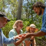 Four adults outdoors in a forested area observing and holding a small object together, smiling and engaged in conversation.