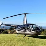 A silver and black Robinson R44 helicopter with registration VH-NSB is parked on a grassy field under a clear blue sky.