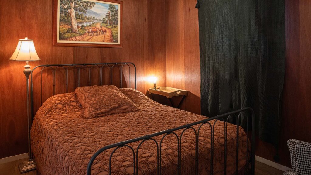 A bedroom with wood-paneled walls, a metal bed frame with a brown textured bedspread, a lamp, a small wall-mounted shelf, and a window covered by a dark curtain.