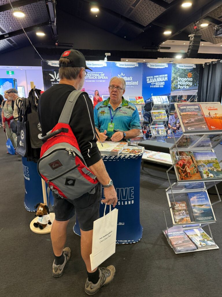 Two men stand at a tourism booth with brochures and maps; one wears a blue shirt behind the table, the other has a backpack and shopping bag, and display racks are visible in the background.