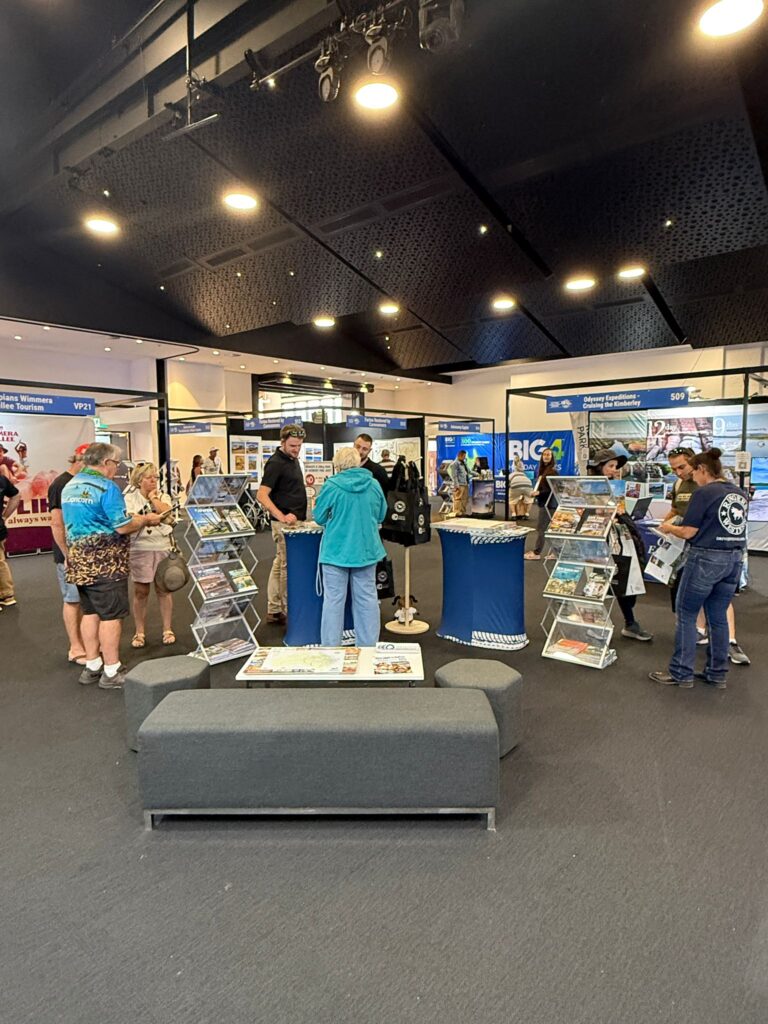 People browse pamphlets and displays at an indoor exhibition or trade show with informational booths and posters visible in the background.