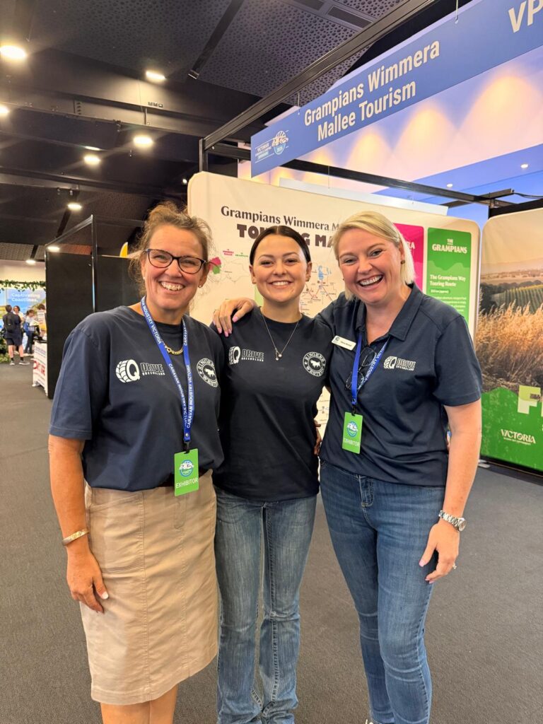 Three women wearing matching navy shirts and name badges stand together smiling at an indoor tourism event, with booths and banners visible in the background.