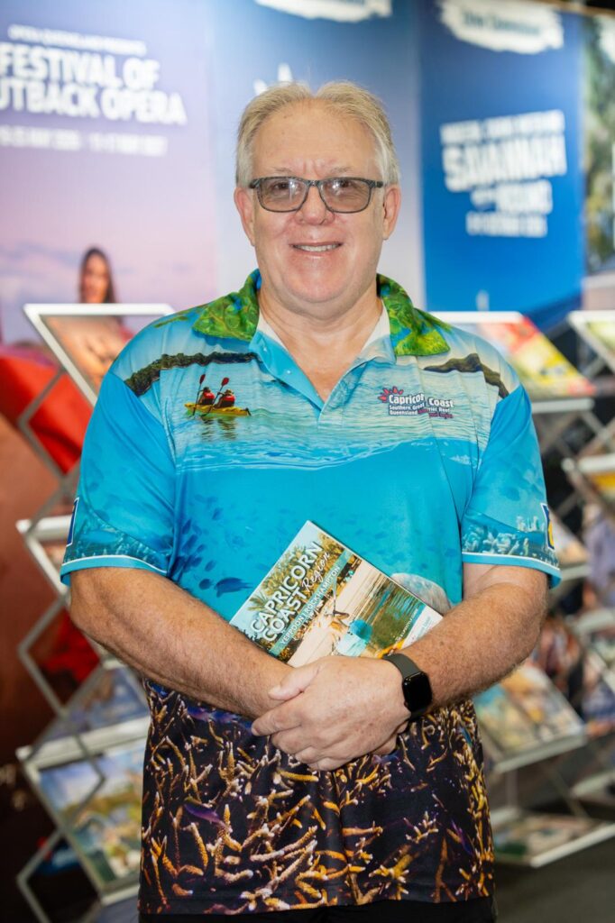 A man wearing glasses and a colorful shirt stands indoors, smiling and holding a tourism brochure. Display racks with brochures are visible in the background.