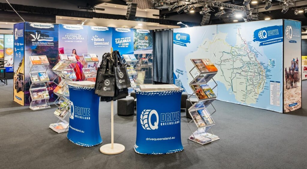 Exhibition booth for Drive Queensland featuring brochures, maps, display stands, promotional bags, and a large map of Queensland on the wall.