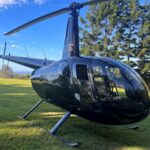 A black helicopter is parked on a grassy field with trees and a blue sky in the background.