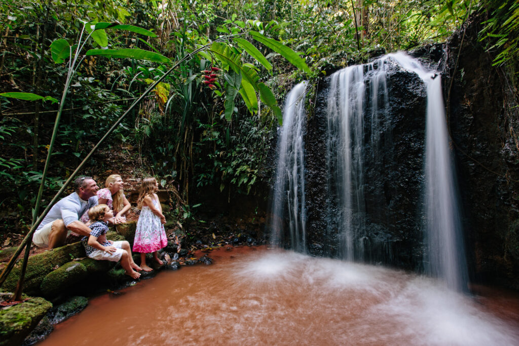 A family sits near a small waterfall in a lush, green forest, with three children observing the water and parents seated beside them.