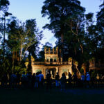 A group of people stands in front of an ornate, castle-like structure surrounded by dense trees at dusk.