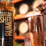 A close-up of a Sugar Shed Rum bottle next to a copper cup, with a hand pouring liquid into the cup. Wooden barrels are blurred in the background.