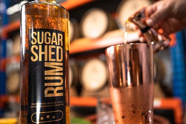 A close-up of a Sugar Shed Rum bottle next to a copper cup, with a hand pouring liquid into the cup. Wooden barrels are blurred in the background.