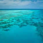 Aerial view of a boat floating on clear turquoise water above coral reefs, with small patches of darker coral visible beneath the surface.