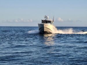 A white motorboat moves quickly across blue open water, creating waves and white spray behind it under a clear sky.