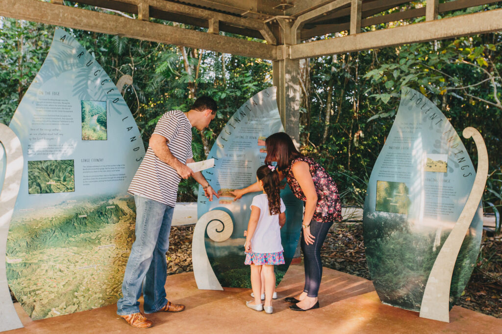 Three people, two adults and one child, read and point at informational display panels in an outdoor covered area surrounded by trees.