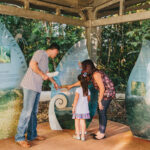 Three people, two adults and one child, read and point at informational display panels in an outdoor covered area surrounded by trees.