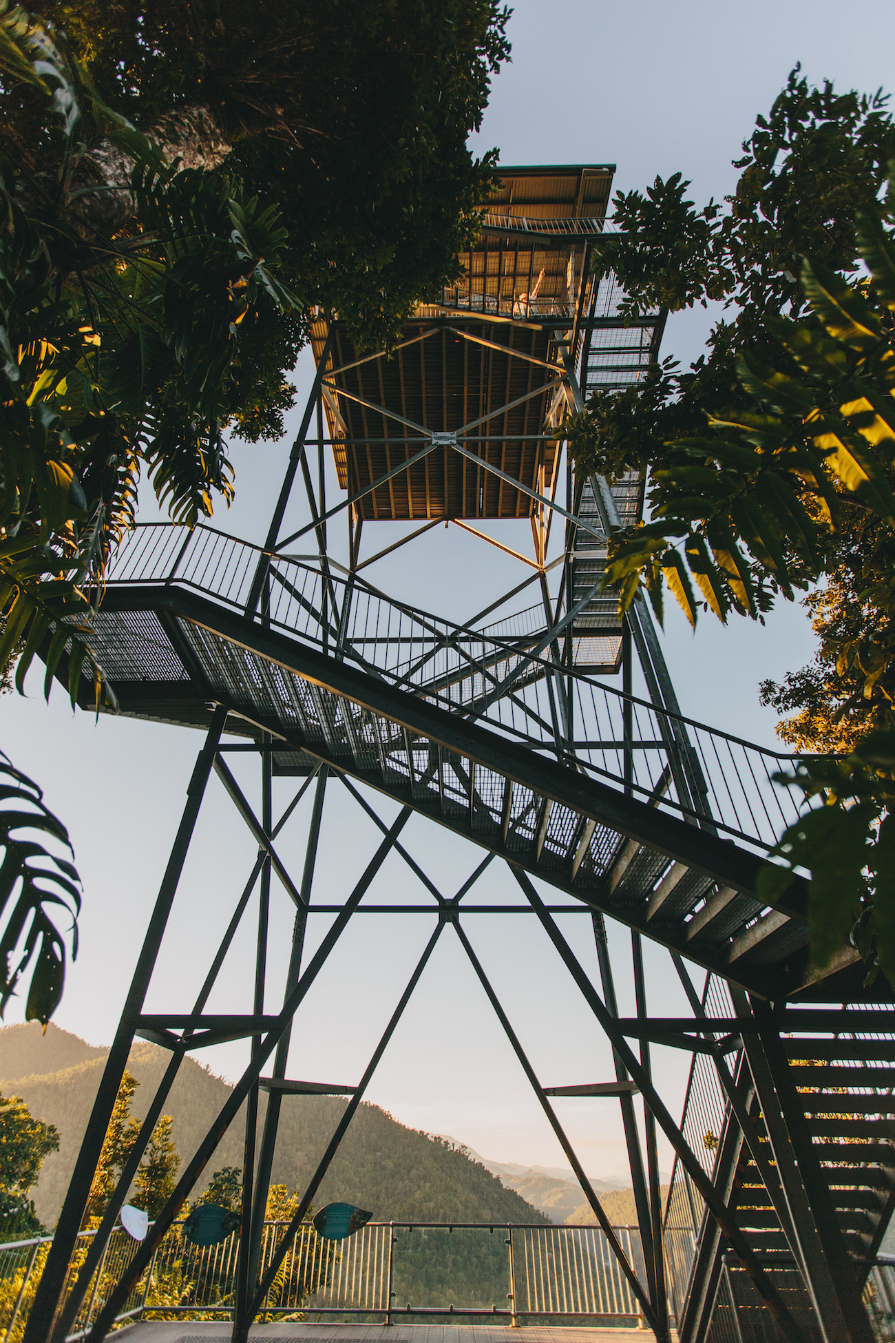 Tall metal observation tower with staircases, surrounded by trees, and overlooking a distant mountain landscape under a clear sky.
