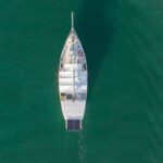 Aerial view of a white sailboat with solar panels floating on calm green water, docked near other boats.
