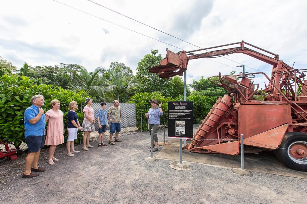A group of people listens to a guide explaining an old red farming machine displayed outdoors next to an informational sign.
