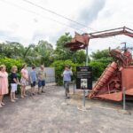 A group of people listens to a guide explaining an old red farming machine displayed outdoors next to an informational sign.