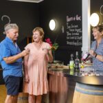 Two older adults sample drinks at a bar while a bartender pours liquor. Chalkboards with cocktail recipes and a quiz are visible in the background.