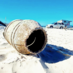 A large rusty metal drum lies on a sandy beach with a white vehicle and the ocean in the background under a clear blue sky.