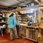 A person stands at a rustic wooden bar counter while an employee behind the counter points at items on a shelf; shelves display jars, bottles, and decor.
