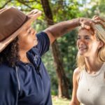 A woman in a hat applies orange paint to another woman's face outdoors in a forested area.