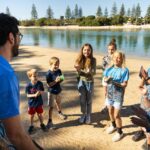 A group of children and an adult stand in a circle on a sandy riverbank, smiling and clapping during an outdoor activity on a sunny day.