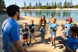 A group of children and an adult stand in a circle on a sandy riverbank, smiling and clapping during an outdoor activity on a sunny day.