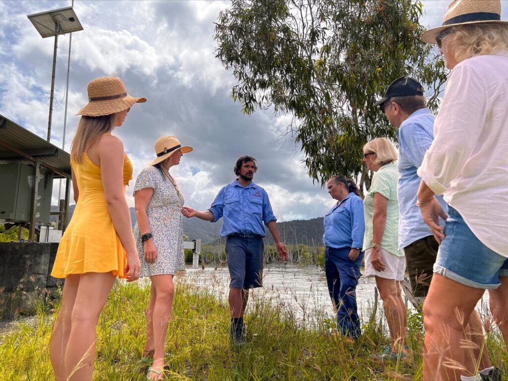 A group of people stand outdoors by a body of water, listening to a man in a blue shirt speaking. Some wear hats and summer clothing; trees and clouds are visible in the background.
