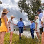 A group of people stand outdoors by a body of water, listening to a man in a blue shirt speaking. Some wear hats and summer clothing; trees and clouds are visible in the background.