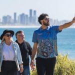 Three people walk along a coastal path with city buildings in the background; one man gestures while talking to the others.