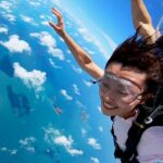 A person wearing goggles and harness is skydiving above a blue ocean with scattered clouds, smiling with arms extended.