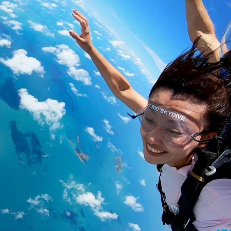 A person wearing goggles and harness is skydiving above a blue ocean with scattered clouds, smiling with arms extended.