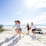 A family of four walks up a sandy dune at the beach on a sunny day, with a van and surfboards parked in the background near the ocean.