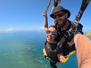 Two people tandem skydiving high above a clear blue ocean, smiling at the camera, with islands visible in the distance.