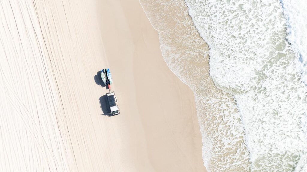 Aerial view of a sandy beach with a white vehicle parked near the water; ocean waves approach the shore from the right.