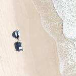 Aerial view of a sandy beach with a white vehicle parked near the water; ocean waves approach the shore from the right.