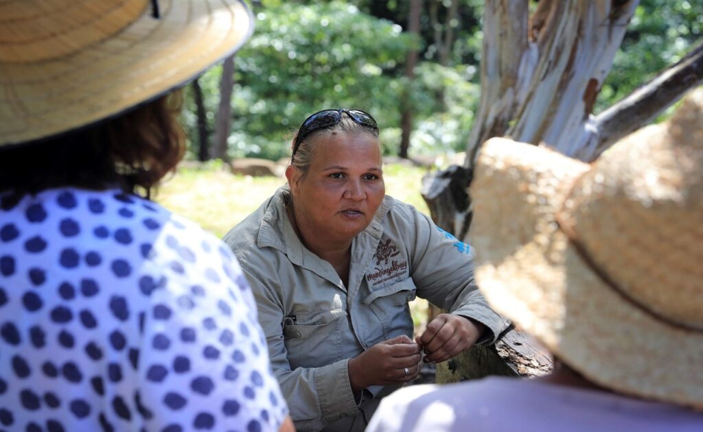 A woman in a tan shirt and sunglasses on her head speaks to two people in hats outdoors, with trees in the background.