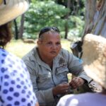 A woman in a tan shirt and sunglasses on her head speaks to two people in hats outdoors, with trees in the background.