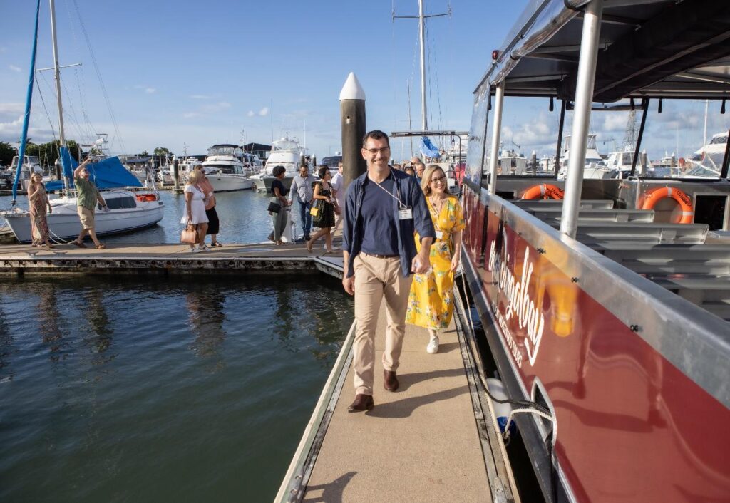People walk off a red boat onto a dock at a marina, with sailboats and other boats in the background on a sunny day.