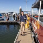 People walk off a red boat onto a dock at a marina, with sailboats and other boats in the background on a sunny day.